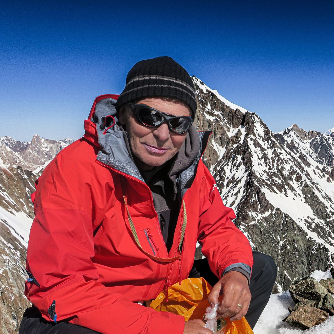 Simon Yates - Man wearing red coat, black beanie hat and sunglasses sat on a snowy mountain.
