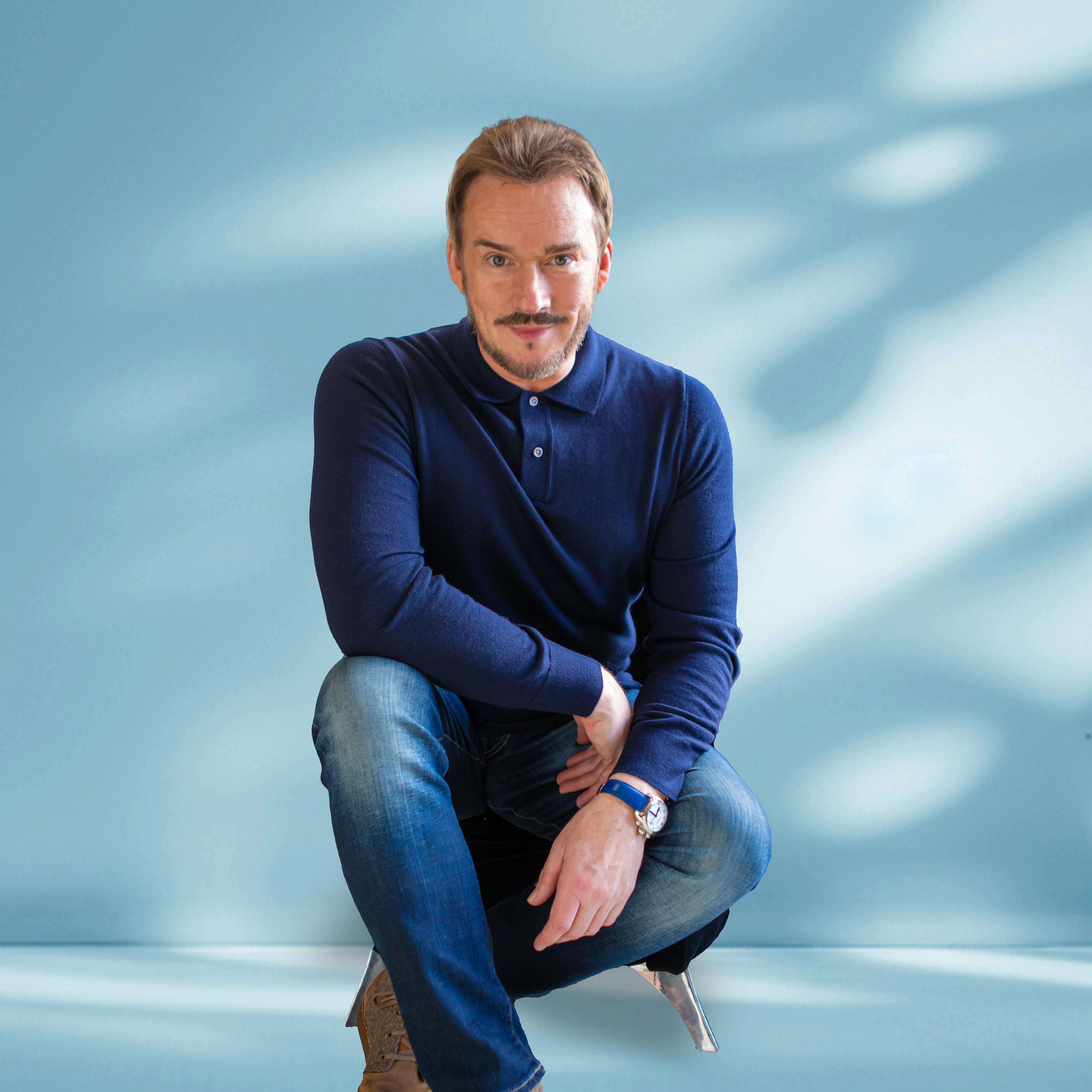 Russell Watson (male) in dark navy blue jumper crouching down with a light blue background behind him